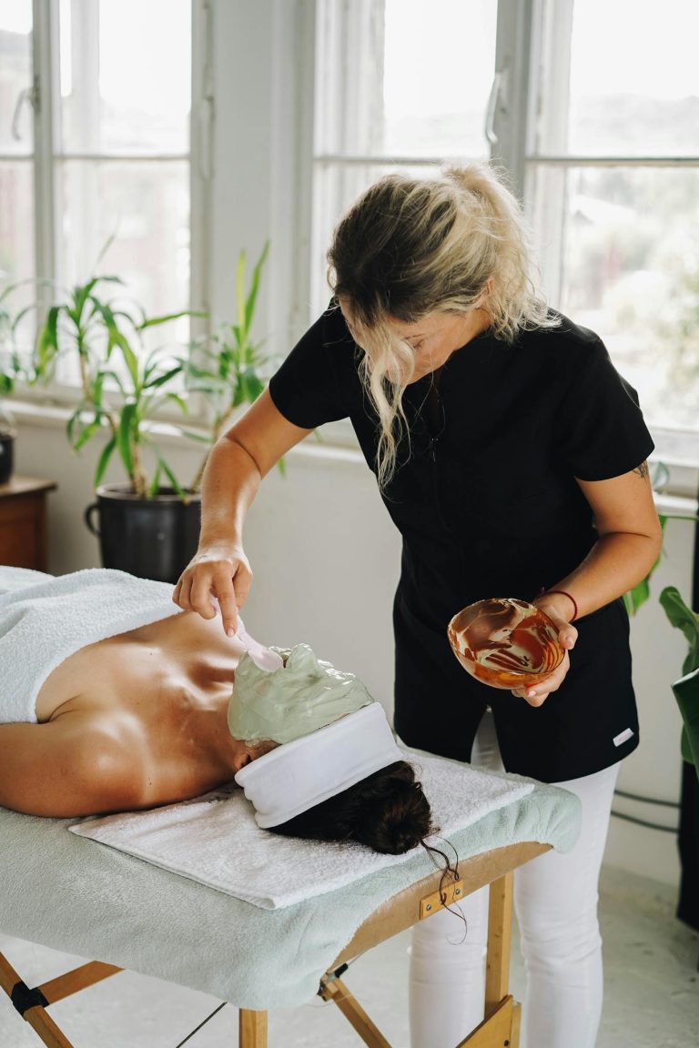 A woman receives a rejuvenating clay facial mask at a spa, surrounded by lush indoor plants.