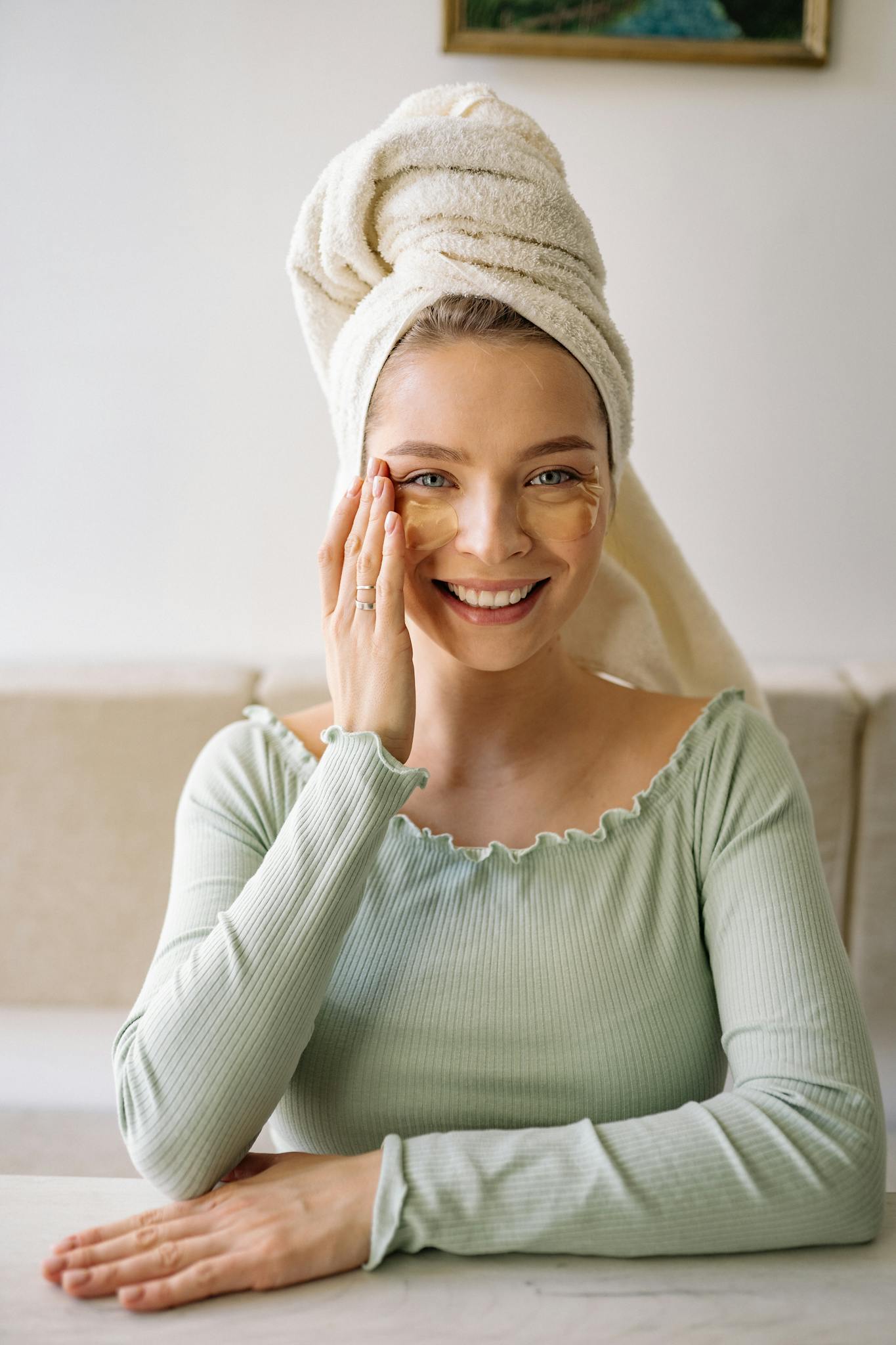 Smiling woman with eye patches and towel on head enjoying a beauty skincare routine indoors.